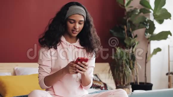 快乐的非裔美国女孩聊天和短信智能手机坐在床上在家里年轻女子视频的预览图