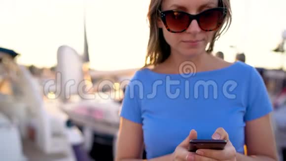 戴墨镜的女人在智能手机上说话在日落时分在码头上和很多游艇和船一起走很近视频的预览图