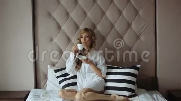 早晨金发姑娘穿着白色衬衫坐在窗边的床上视频的预览图