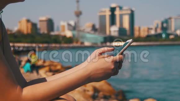 年轻女性使用移动电话坐在海边的堤岸和梅格波利斯摩天大楼上视频的预览图