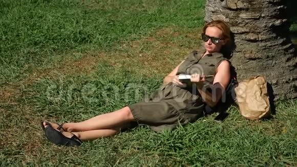 金发女人在公园里看书坐在草地上树下4k慢镜头视频的预览图