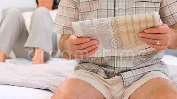 成熟的男人看报纸而他的妻子听音乐视频的预览图