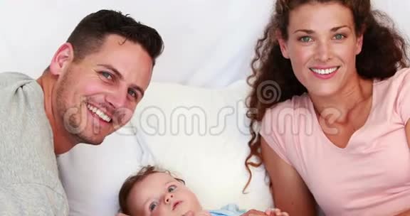 穿着蓝色婴儿裤的小男孩被快乐的父母在床上挠痒痒视频的预览图