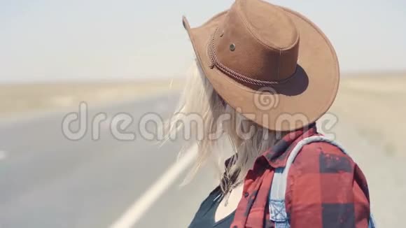年轻时尚的女孩穿着牛仔衣服在沙漠里搭便车在路上她背着背包和帽子通行证视频的预览图