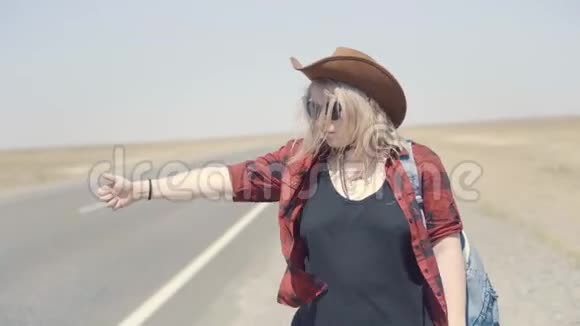 年轻时尚的女孩穿着牛仔衣服在沙漠里搭便车在路上她背着背包和帽子通行证视频的预览图