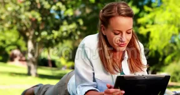 一个漂亮的金发美女在公园里用平板电脑躺在毯子上视频的预览图