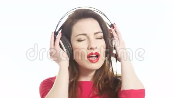 漂亮的红色女孩随着音乐在大耳机中摇摆注意到相机和白色背景上的隔离视频的预览图