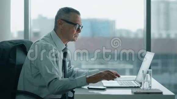 老商人在现代办公室里看电话和电脑视频的预览图