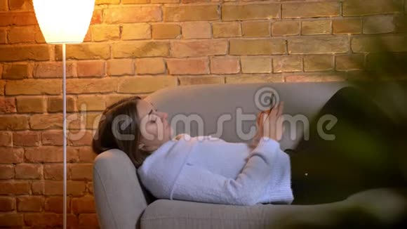 年轻美丽的白种人女性躺在沙发上在舒适的室内家里发短信特写照片视频的预览图