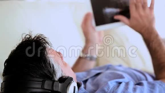 成熟的男人躺在沙发上听音乐用平板电脑视频的预览图