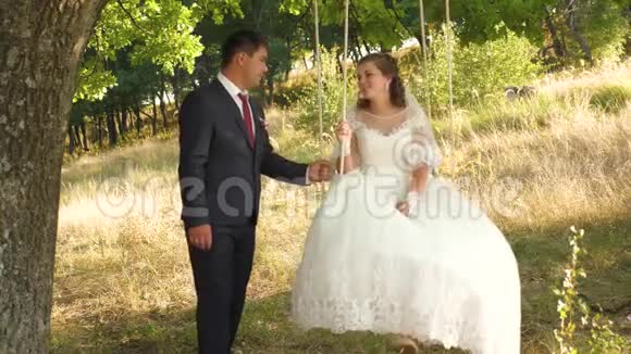 快乐的新娘和新郎穿着漂亮的白色连衣裙在夏季公园的秋千上摇摆夏天在橡树枝上荡秋千视频的预览图