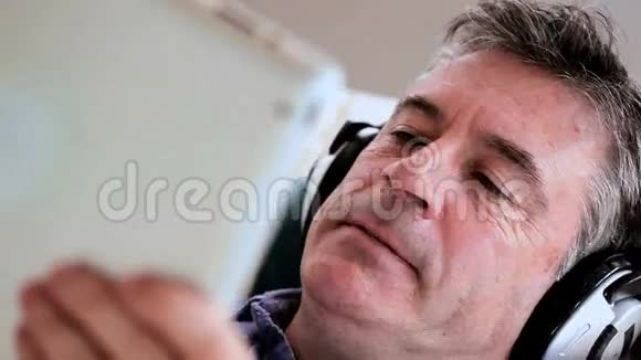 成熟的男人躺在沙发上用他的数码平板电脑听音乐视频的预览图