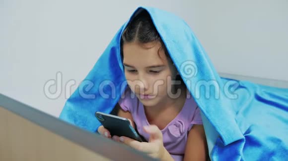 带智能手机的毯子下可爱女孩的肖像小女孩在社交媒体毯子下玩网络游戏女孩视频的预览图