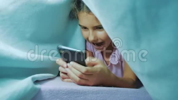 带智能手机的毯子下可爱女孩的肖像在社交媒体下玩网络游戏的小女孩生活方式视频的预览图