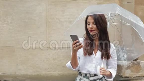 美丽的年轻商务女性在雨天的街道上使用智能手机微笑拿着雨伞交流视频的预览图