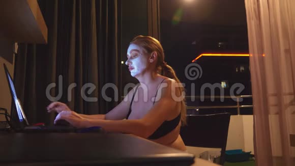 晚上带着化妆面具的年轻女人在家里用笔记本电脑工作自然视频的预览图