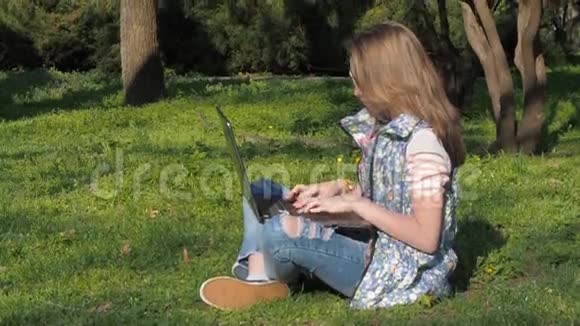 一个十几岁的女孩在公园里带着笔记本电脑漂亮的女孩正拿着笔记本电脑坐在草地上春天阳光明媚的一天视频的预览图
