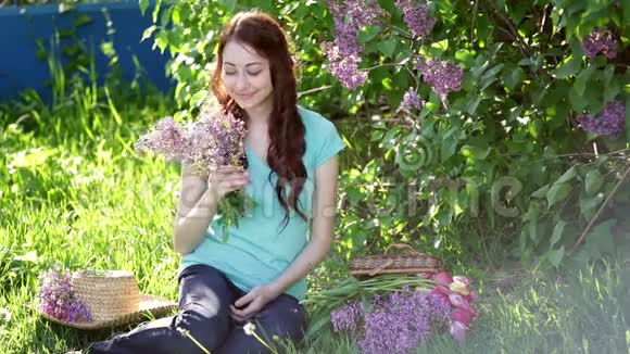 美丽的女人带着一束紫丁香坐在草地上的春天公园里视频的预览图