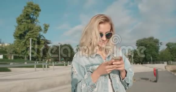 阳光明媚的一天年轻的金发女人戴着太阳镜在城市里用手机视频的预览图