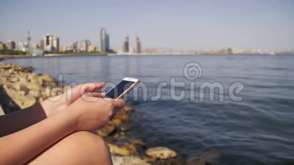 年轻女性使用智能手机坐在海边的堤岸和梅格波利斯摩天大楼上视频的预览图