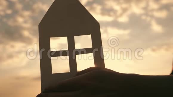 日落时分在家中的田野里双手紧握着纸模纸屋的剪影在光线下视频的预览图