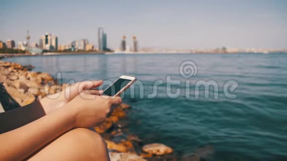 年轻女子在海边的堤岸上坐着手机高楼大厦林立视频的预览图