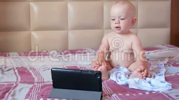 可爱的孩子坐在床上盯着平板电脑上的卡通一岁的小婴儿没有衣服视频的预览图