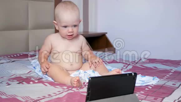 可爱的孩子坐在床上盯着平板电脑上的卡通一岁的小婴儿没有衣服视频的预览图