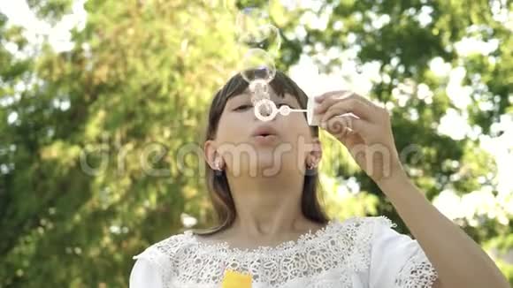 小女孩在公园里玩把泡泡吹进镜头慢动作漂亮的女孩吹肥皂视频的预览图