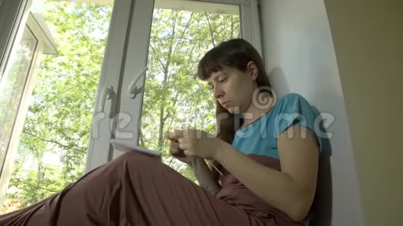 年轻女子用坐在窗台上的针织针编织蓝色的纱线视频的预览图