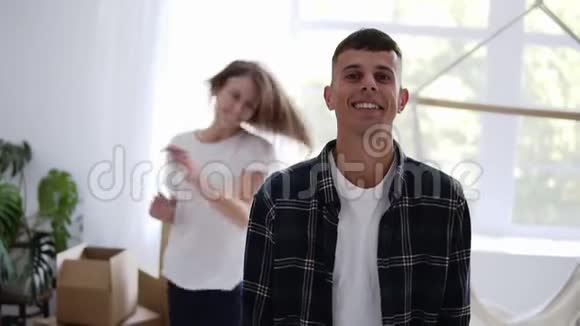 快乐的白种人男性在新公寓里缓慢地微笑而他的女朋友在背景上跳舞然后跳到他的身上视频的预览图