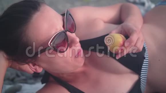 一个穿泳衣的女孩躺在海滩上吃桃子的脸的特写一个女孩吃一个美味的桃子视频的预览图
