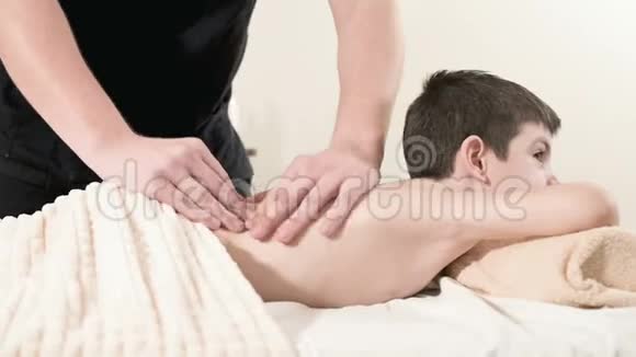 一位男性理疗师按摩师为躺在按摩床上的一个微笑的小男孩做一个疗愈放松的按摩按摩视频的预览图