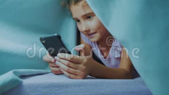带智能手机的毯子下可爱女孩的肖像在社交媒体下玩网络游戏的小生活方式女孩视频的预览图