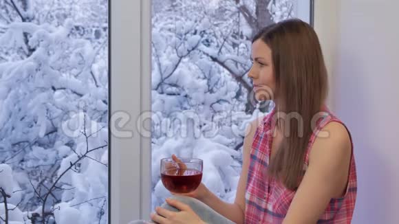 可爱的女孩坐在窗台上看着窗外喝茶冬天在外面视频的预览图