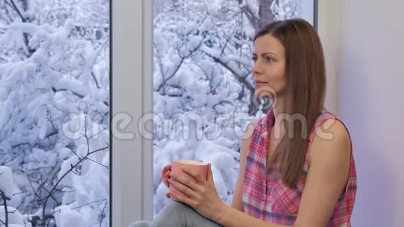 漂亮的女孩坐在窗台上看着窗户喝着茶冬天在外面视频的预览图