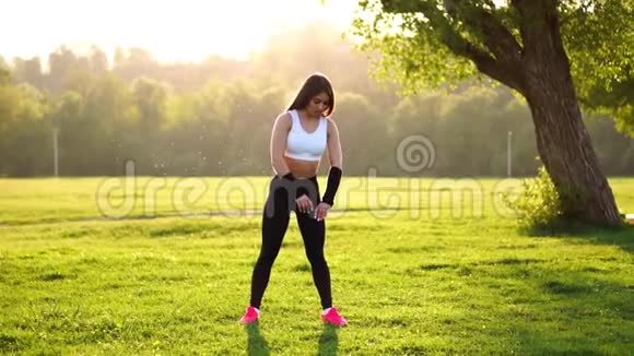 穿着白色上衣黑色紧身裤和粉色运动鞋的运动型黑发女郎在阳光下伸展肌肉准备视频的预览图