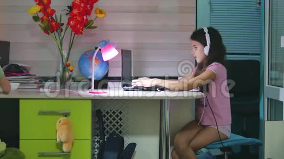 带着耳机坐在室内的十几岁女孩带着笔记本电脑在笔记本电脑上玩网络游戏的女生视频的预览图