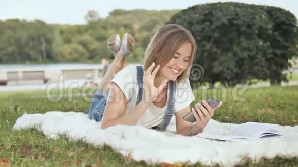一个年轻的女孩躺在公园里的白色格子布上在电话里拨了一条信息视频的预览图
