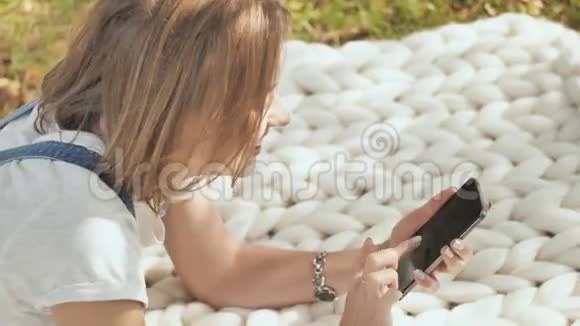 一个年轻的女孩躺在公园里的白色格子布上在电话里拨了一条信息视频的预览图