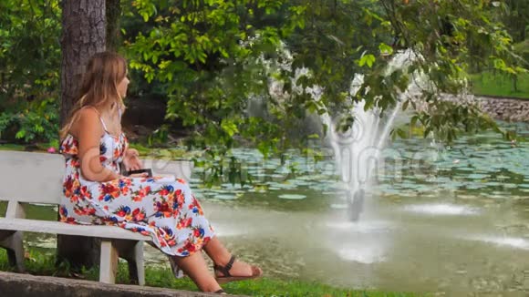 金发女郎坐在喷泉旁的长凳上公园里有iPhone视频的预览图