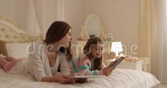 母亲与女儿躺在床上使用平板电脑快乐的年轻家庭视频的预览图
