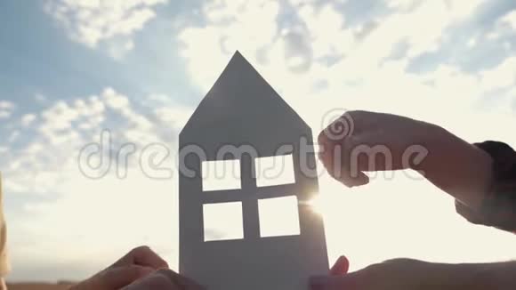 日落时分在家中的田野里双手紧握着纸模纸屋的剪影在光线下视频的预览图