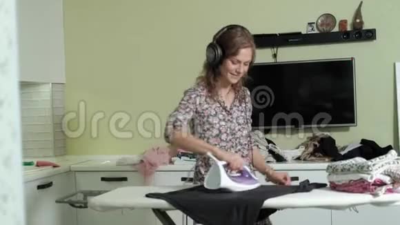 女人在家里熨衣服在厨房里听着耳机上的音乐和跳舞视频的预览图