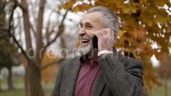 一个戴眼镜用电话的英俊的老人秋天在公园里散步视频的预览图