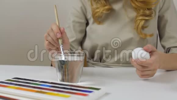 面目全非的女人用刷子涂复活节彩蛋桌子上有一杯水和一碗鸡蛋筹备视频的预览图