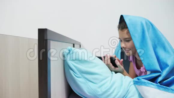 十几岁的女孩在毯子下玩智能手机睡前的小女孩躺在床上上网视频的预览图