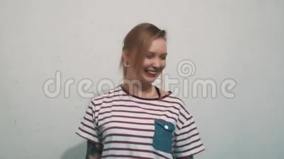 穿着条纹衬衫的快乐金发女孩纹身在白墙前笑视频的预览图