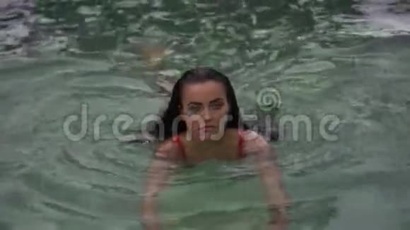 美丽的黑发女人穿着粉红色比基尼在热带度假胜地的室外游泳池放松视频的预览图