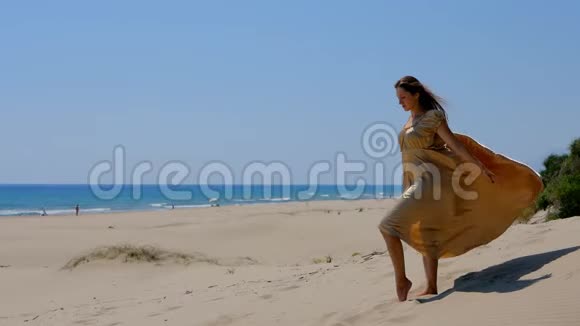 年轻漂亮的女人穿着金色的长裙站在靠近大海的沙丘上视频的预览图